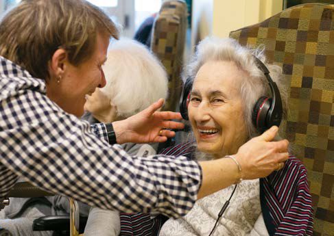 An elderly woman with headphones on smiles at the camera, as a nurse places headphone on her head playing music